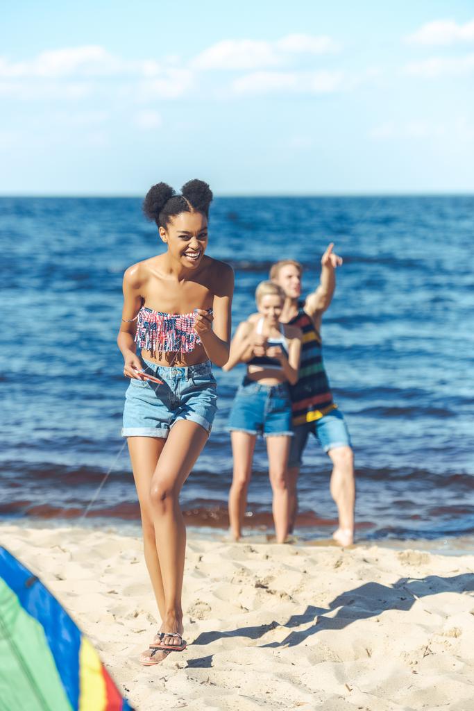 селективное внимание мультикультурных друзей с кайтом, проводящих время на песчаном пляже вместе
 - Фото, изображение