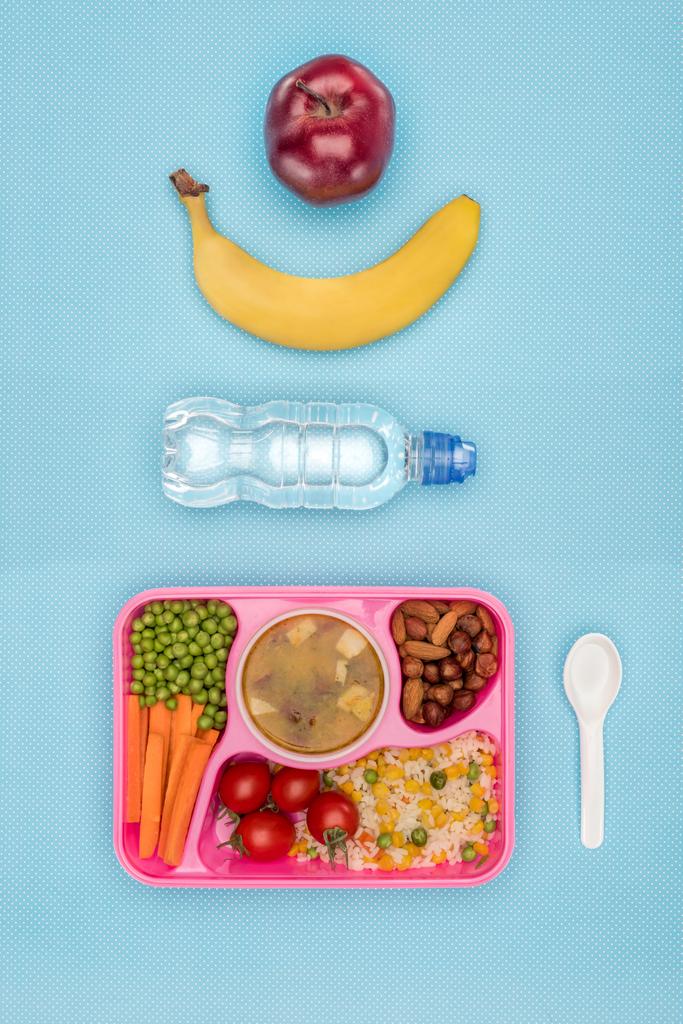 верхний вид подноса с детским обедом для школы, бутылка воды, банан и яблоко на голубой поверхности
 - Фото, изображение