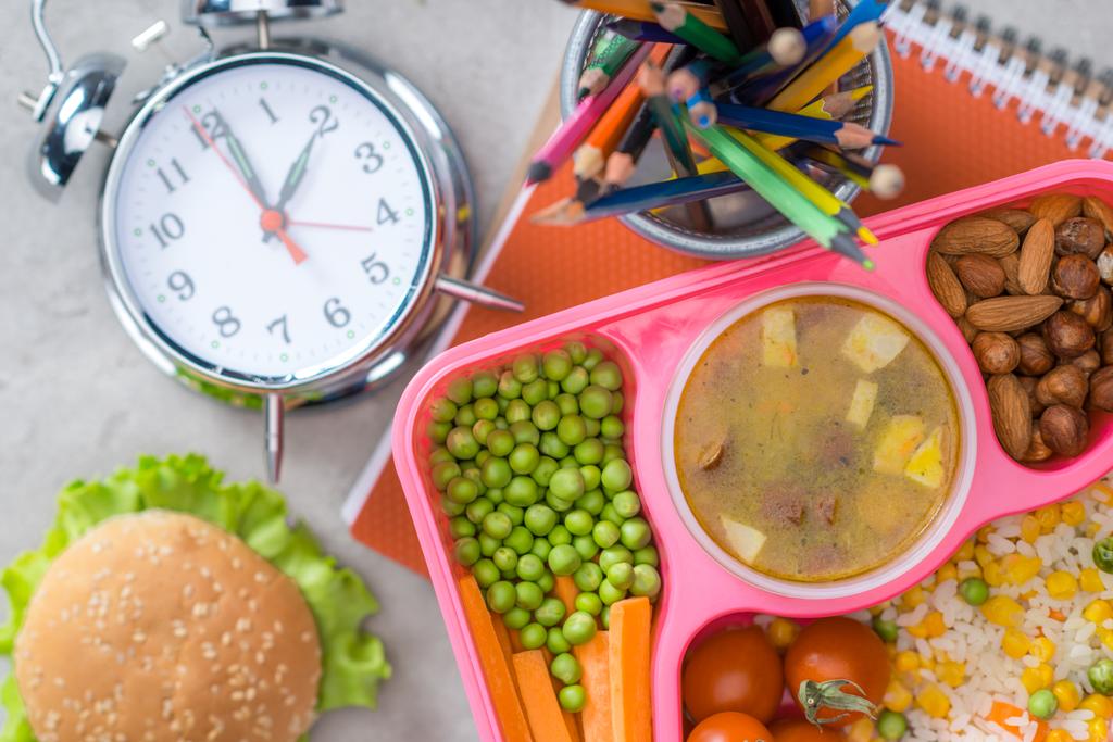 κάτοψη της θήκης με το μεσημεριανό γεύμα τα παιδιά για το σχολείο, Ξυπνητήρι και χρωματιστά μολύβια στο τραπέζι - Φωτογραφία, εικόνα