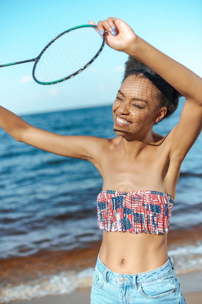 portrait of smiling african american woman with badminton racquet on beach - Photo, Image