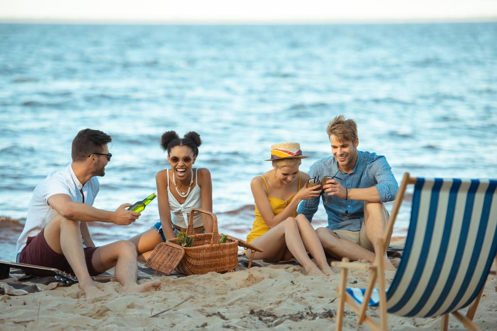 διαφυλετικο, χαμογελώντας νέους φίλους με τα ποτά που στηρίζεται σε αμμώδη παραλία μαζί σε καλοκαιρινή μέρα - Φωτογραφία, εικόνα