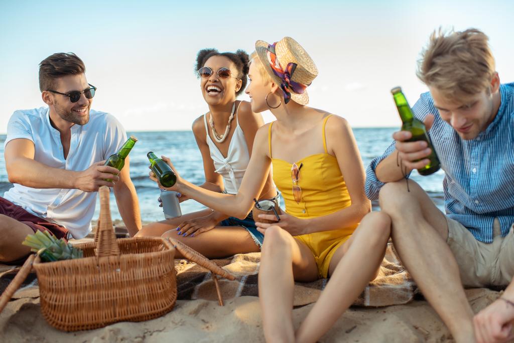 Sex tussen verschillendre rassen jonge vrienden lachen met bier rustend op zandstrand samen op zomerdag - Foto, afbeelding