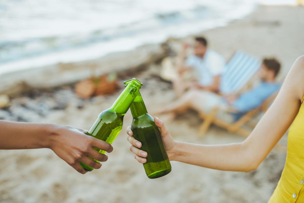selective focus of multiracial friends clinking glass bottles of beer while resting on sandy beach together - Photo, Image