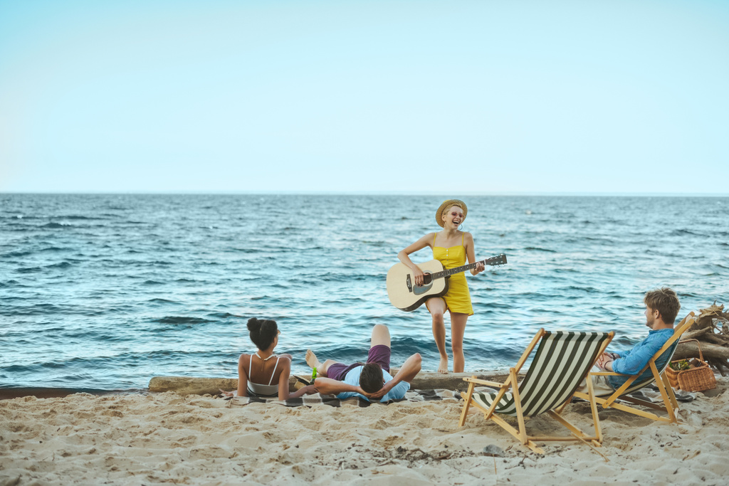 молода жінка грає на акустичній гітарі для багатоетнічних друзів на піщаному пляжі
 - Фото, зображення