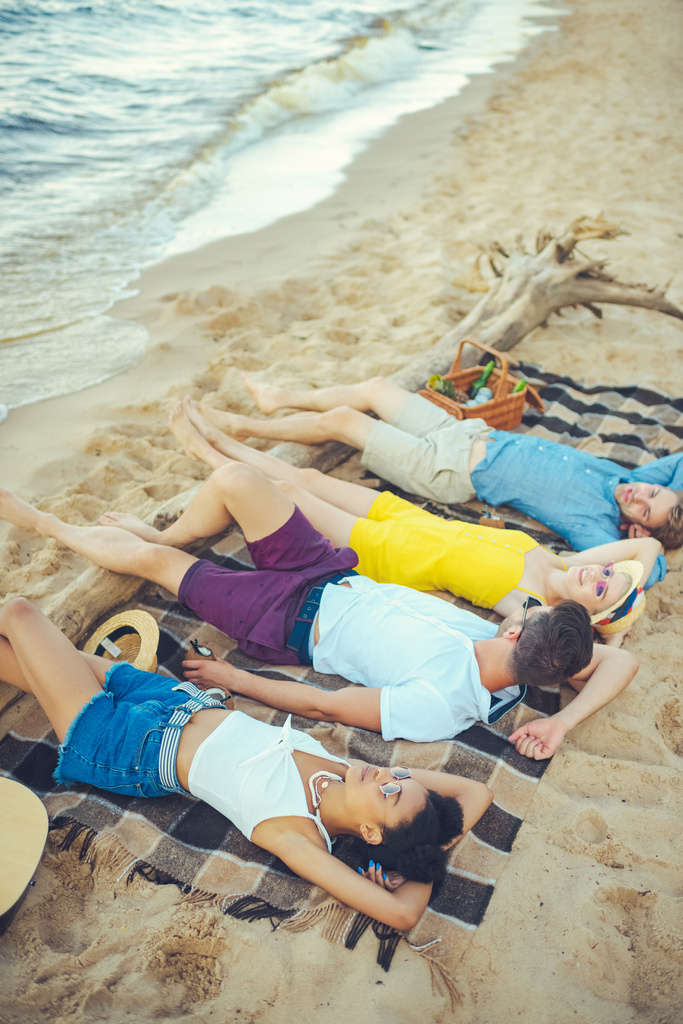 багаторасові молоді люди лежать на ковдрі, проводячи час на піщаному пляжі
 - Фото, зображення