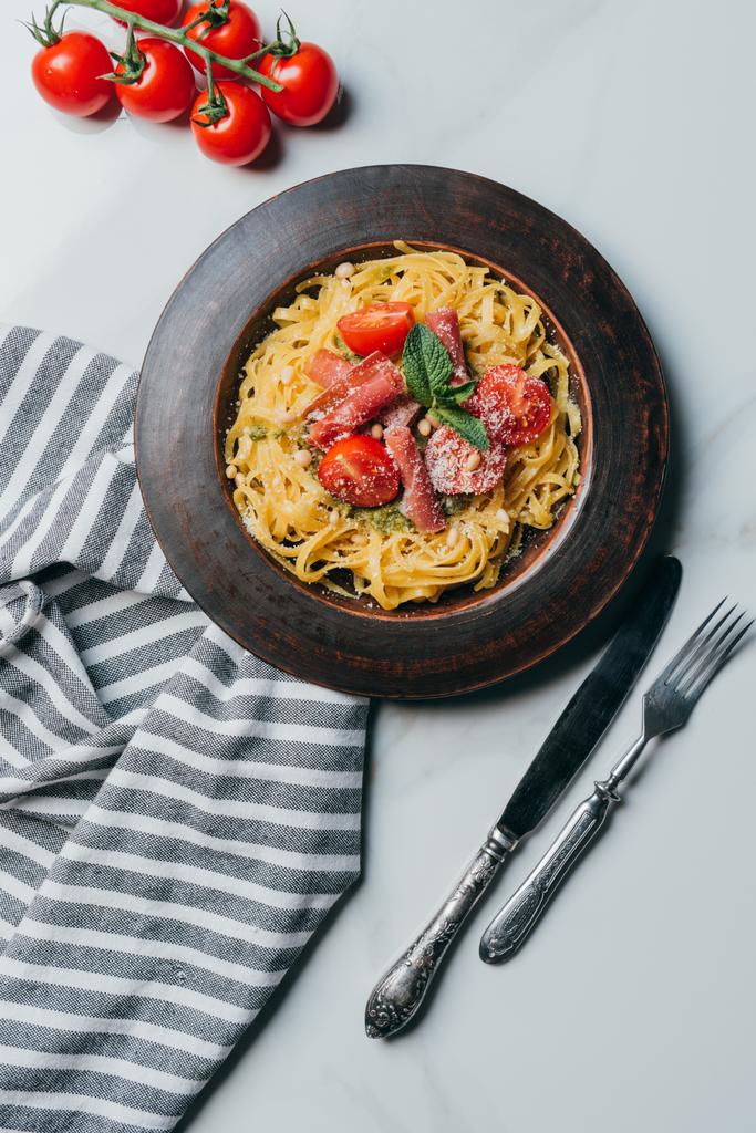 vista superior de la pasta con hojas de menta, jamón y tomates cherry cubiertos por parmesano en el plato en la mesa de mármol con toalla de cocina, cuchillo y tenedor
  - Foto, Imagen