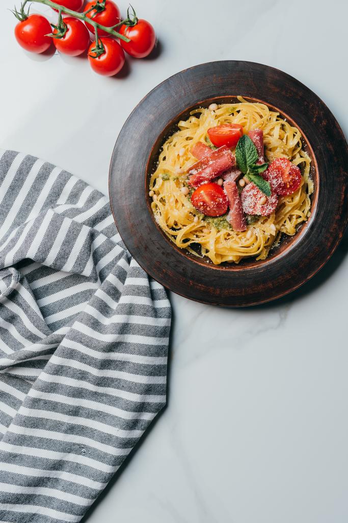 verhoogde weergave van pasta met munt bladeren, Jamón en kerstomaatjes, parmesan vallende op plaat op marmeren tafel met keuken handdoek  - Foto, afbeelding