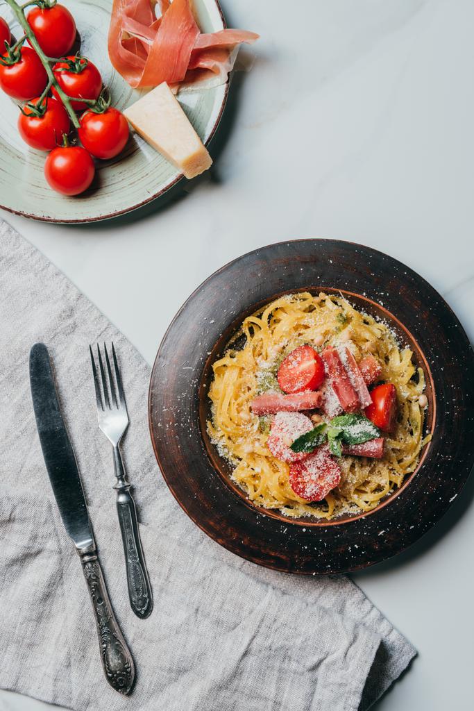 vista elevada de platos con pasta y jamón, tomates parmesano y cereza sobre mesa de mármol con toalla de cocina, tenedor y cuchillo
  - Foto, Imagen