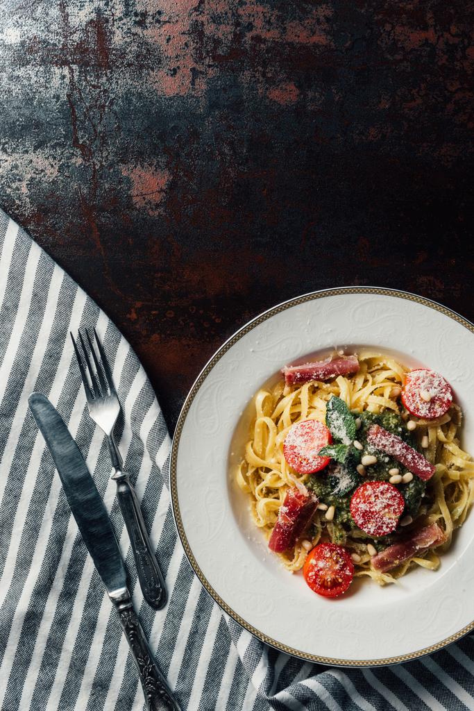 vista elevada de la pasta con hojas de menta, jamón y tomates cherry cubiertos por parmesano rallado en el plato en la mesa con toalla de cocina, cuchillo y tenedor
 - Foto, imagen