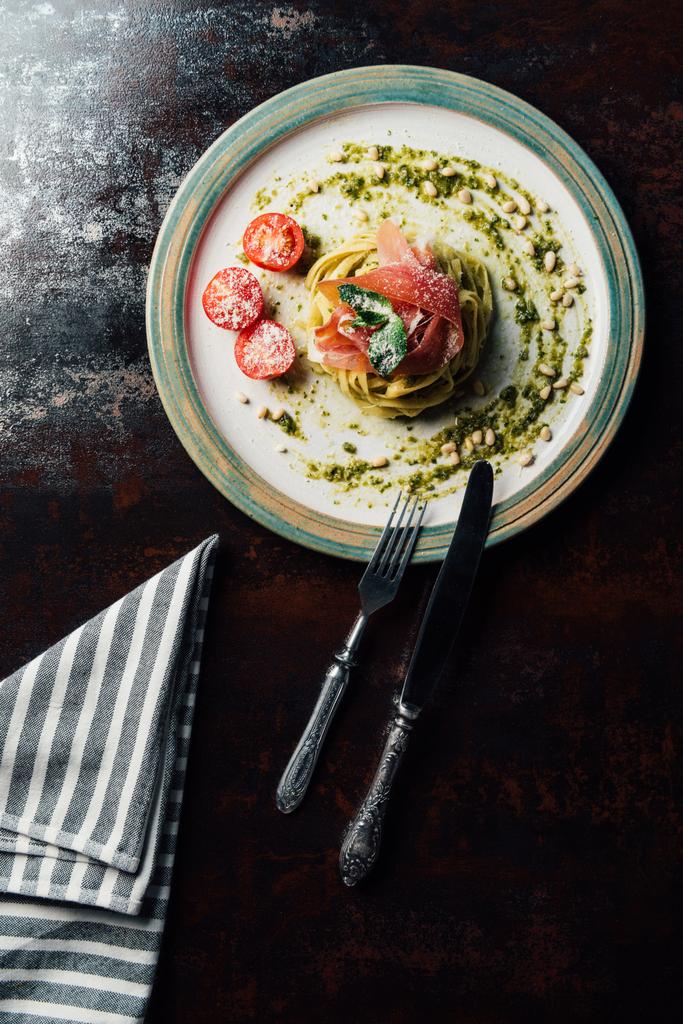 ミントの葉、ハモン、松の実、ペスト板上のテーブル ナイフ、フォーク、キッチン タオルですりおろしたパルメザン チーズで覆われてチェリー トマトとパスタのトップ ビュー  - 写真・画像