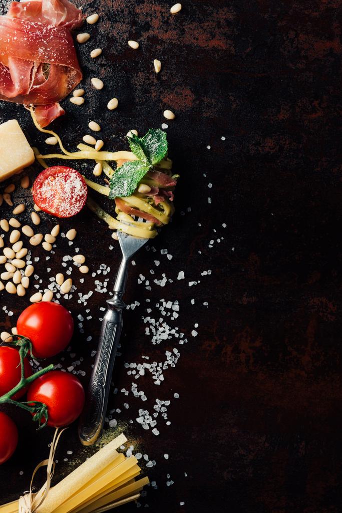 top view of fork wrapped by pasta with mint leaves, jamon, pine nuts,  and cherry tomatoes covered by grated parmesan on table surrounded by ingredients  - Photo, Image