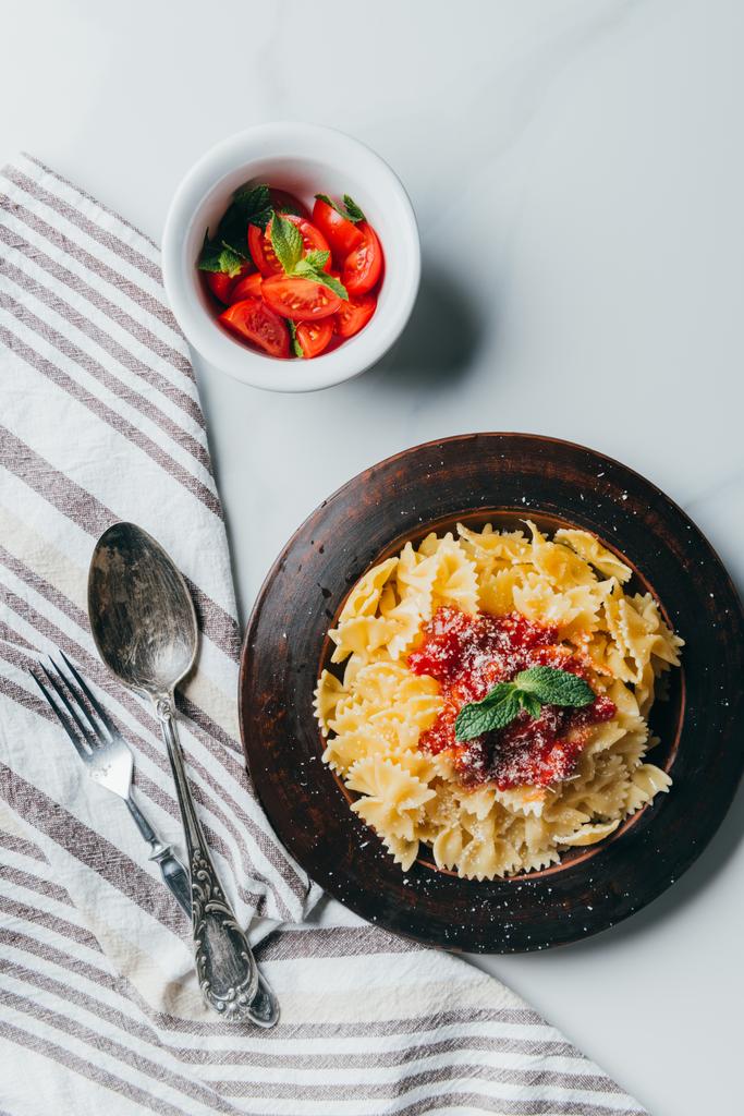 повышенный вид тарелки с макаронами, кухонным полотенцем, вилкой, ложкой и миской с помидорами черри на мраморном столе
  - Фото, изображение