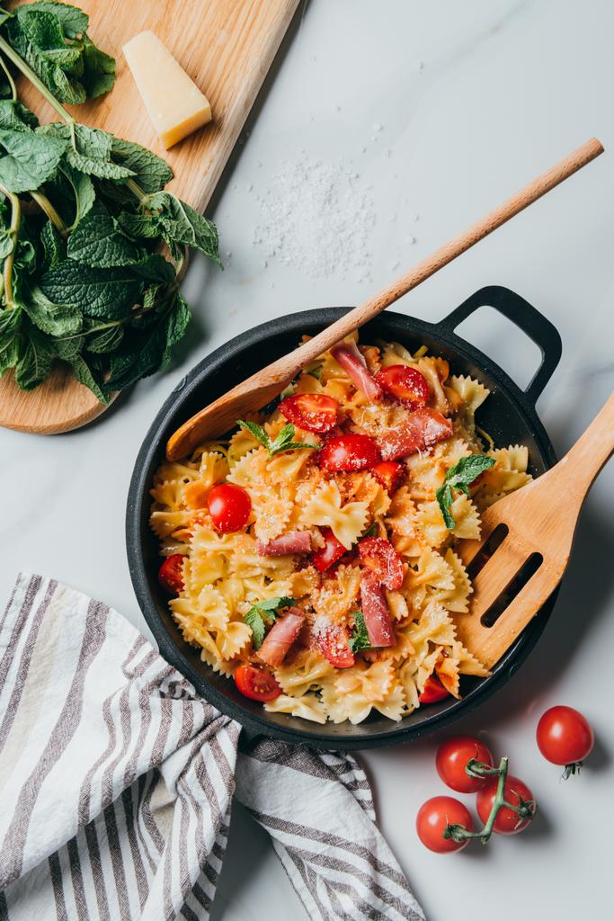 verhoogde weergave van pasta met jamón, cherry tomaten, muntblaadjes gedekt door geraspte Parmezaanse kaas in de pan met houten spatel en lepel op marmeren tafel - Foto, afbeelding