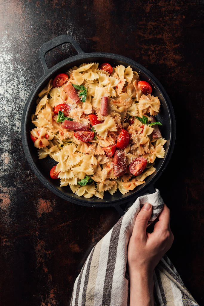 gedeeltelijke weergave van vrouw met pasta met jamón, cherry tomaten, muntblaadjes gedekt door geraspte Parmezaanse kaas in de pan verpakt door keuken handdoek boven tafel  - Foto, afbeelding
