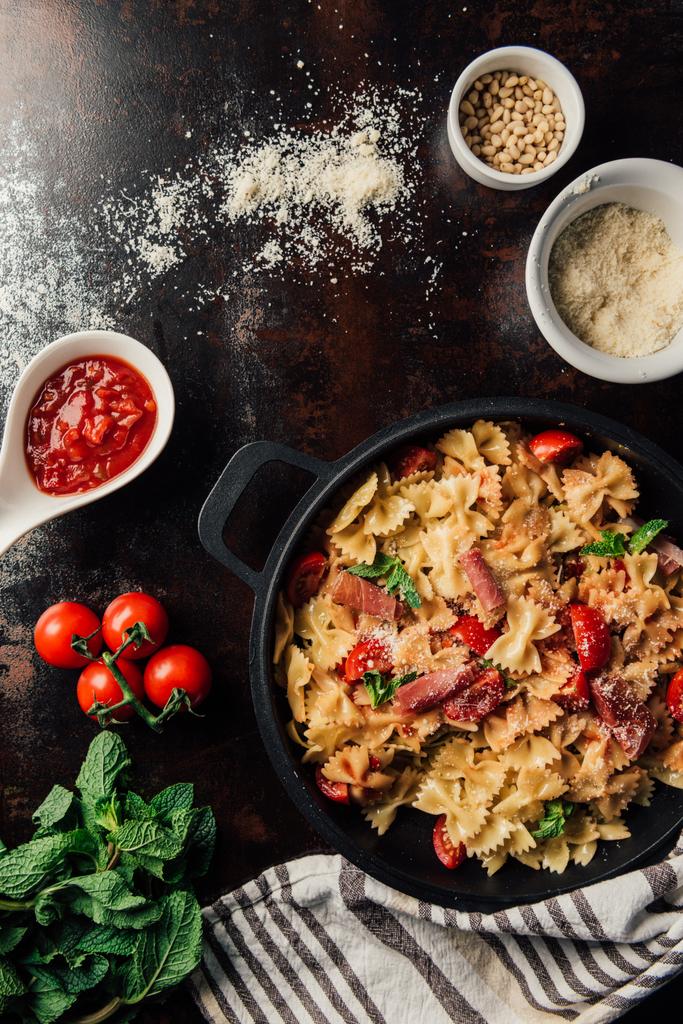 view from above of pasta with jamon, pine nuts, sauce, cherry tomatoes, mint leaves covered by grated parmesan in pan surrounded by ingredients and kitchen towel on table  - Photo, Image