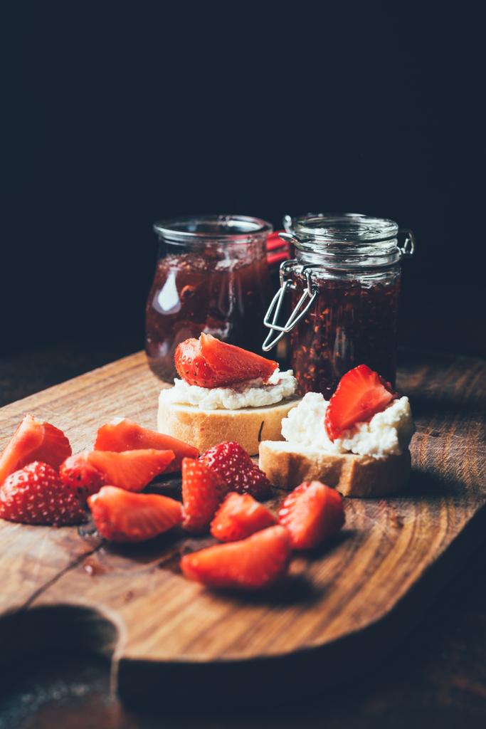 selective focus of sandwiches with cream cheese, strawberry slices and fruit jam on cutting board on black  - Photo, Image