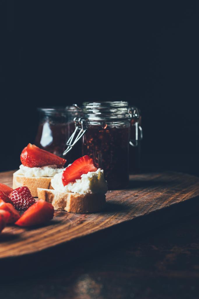 selective focus of sandwiches with cream cheese, strawberry slices and fruit jam on cutting board on black  - Photo, Image