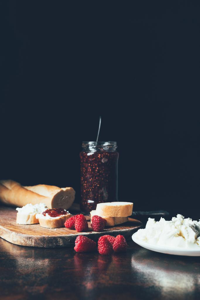 cream cheese in plate, raspberries, jam in jar and baguette on cutting board on black  - Photo, Image