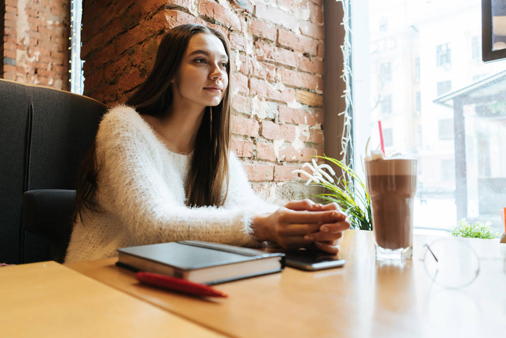hermosa chica de pelo largo en un suéter blanco se sienta en un café bebe delicioso café con leche y espera su comida
 - Foto, Imagen