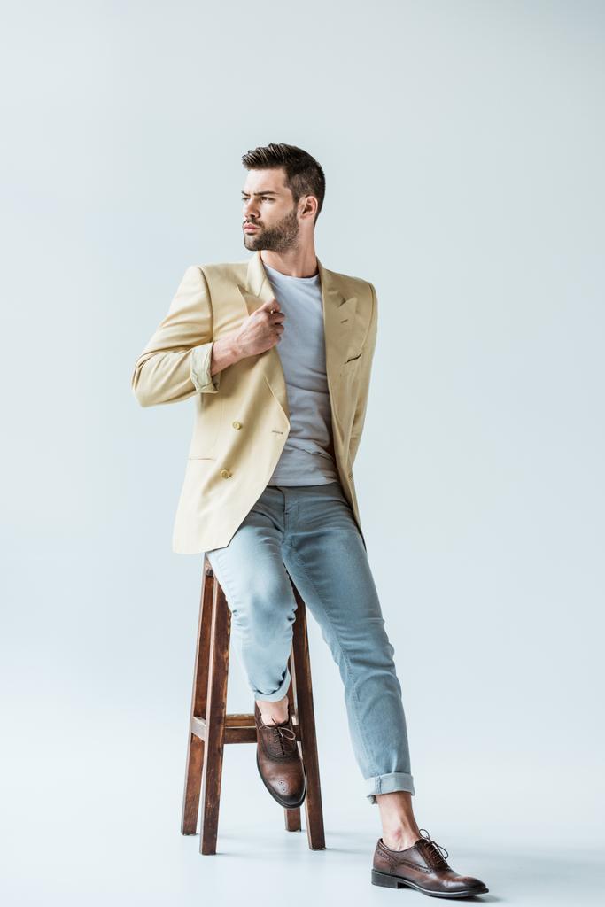 Handsome bearded man in beige jacket sitting on stool on white background - Photo, Image