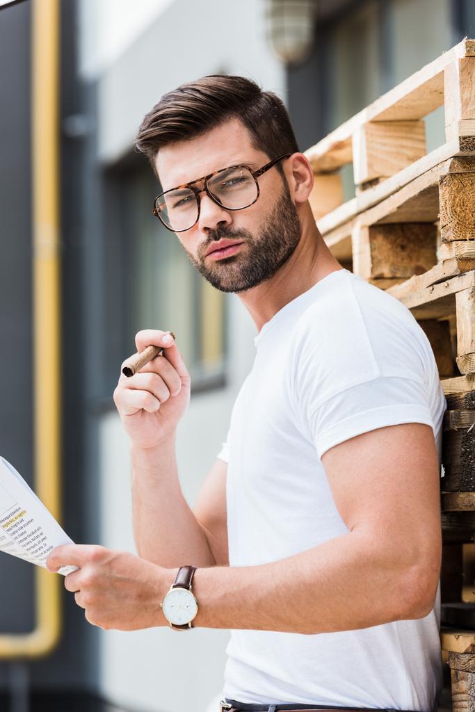 modisch selbstbewusster Geschäftsmann mit Zigarre und Dokument in den Händen, die neben Holzpaletten stehen - Foto, Bild