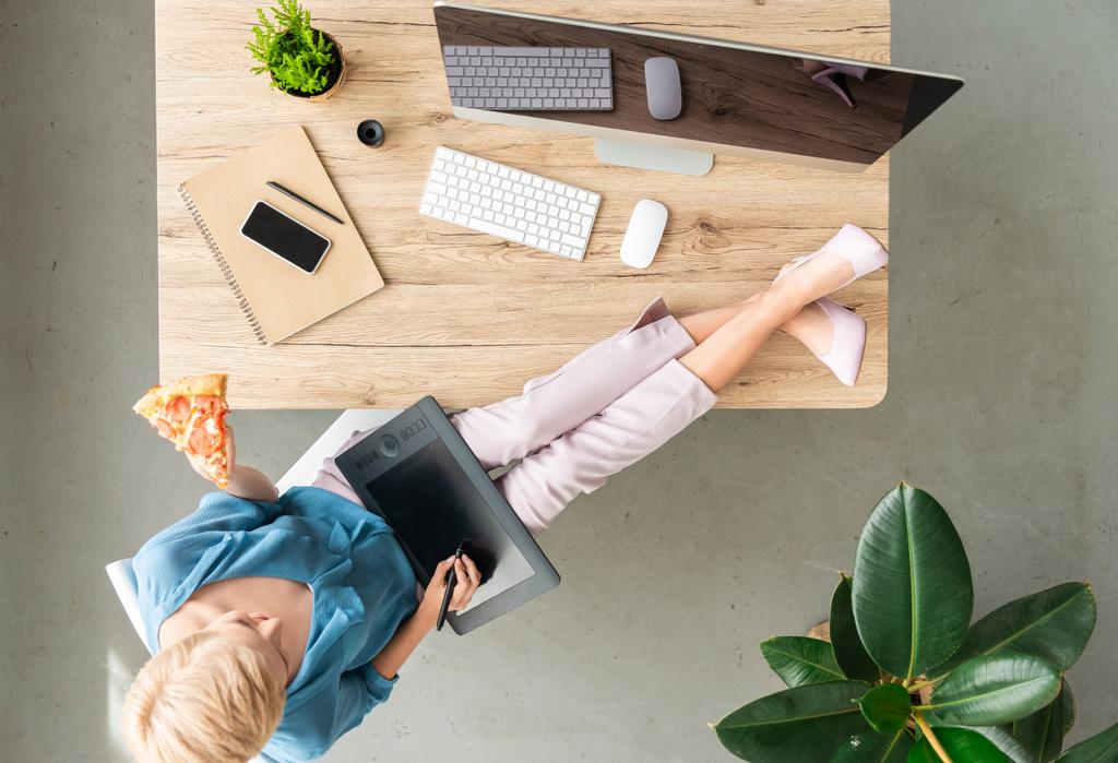 vista superior da elegante freelancer feminino comendo pizza e usando tablet gráfico com pernas na mesa em casa
 - Foto, Imagem