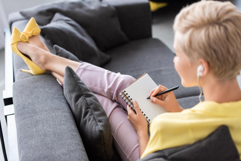 foco seletivo da mulher elegante em fones de ouvido escrevendo no livro didático no sofá em casa
  - Foto, Imagem