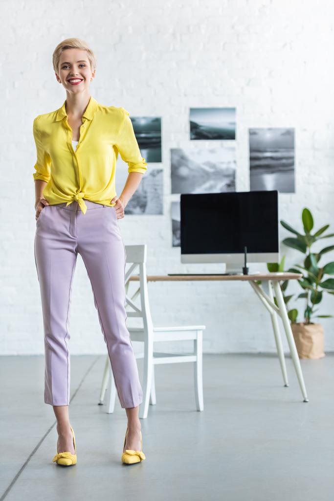 stylish smiling businesswoman with hands in pockets looking at camera at office  - Photo, Image