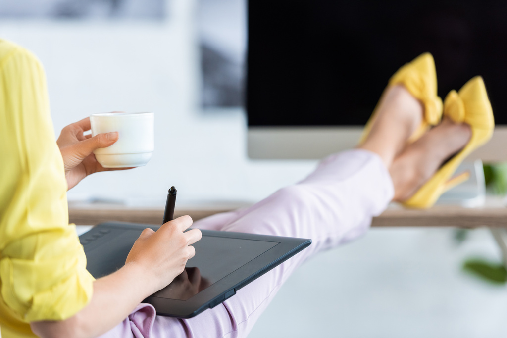 przycięte zdjęcie kobiece freelancer, picia kawy i za pomocą tabletu graficznego przy stole z komputerem w biurze  - Zdjęcie, obraz