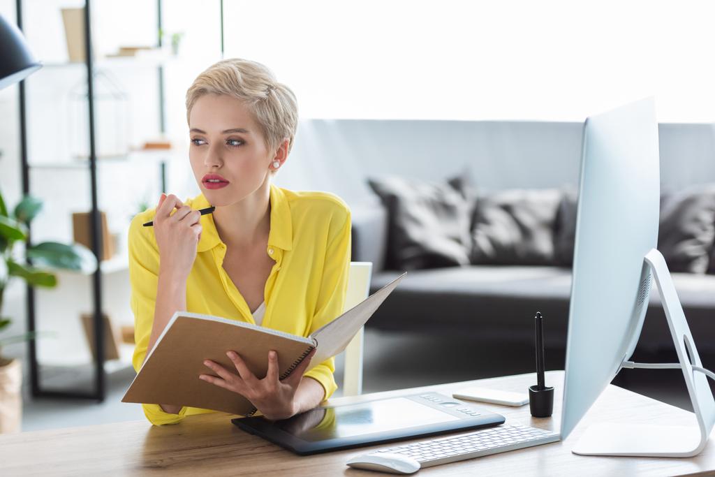 zamyšlená žena na volné noze drží učebnice a sedí u stolu s počítačem a grafický tablet  - Fotografie, Obrázek