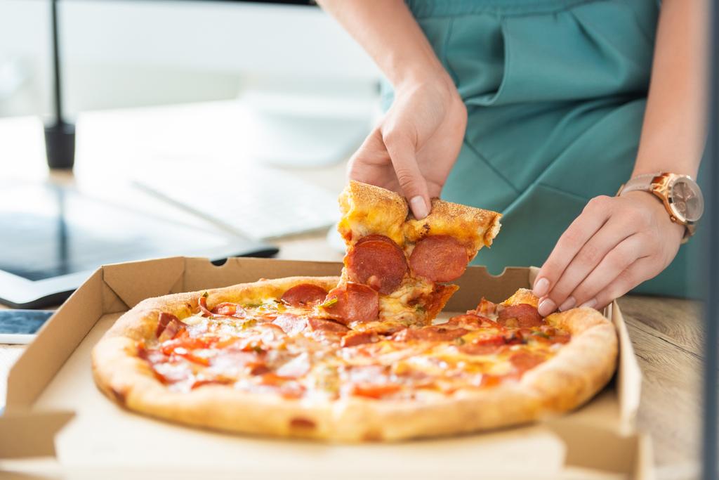 vue partielle d'une pigiste féminine prenant une tranche de pizza à table avec tablette graphique
  - Photo, image