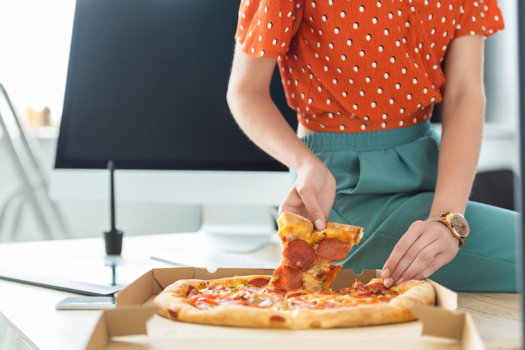 cropped image of female freelancer taking pizza slice from box at table with computer  - Photo, Image
