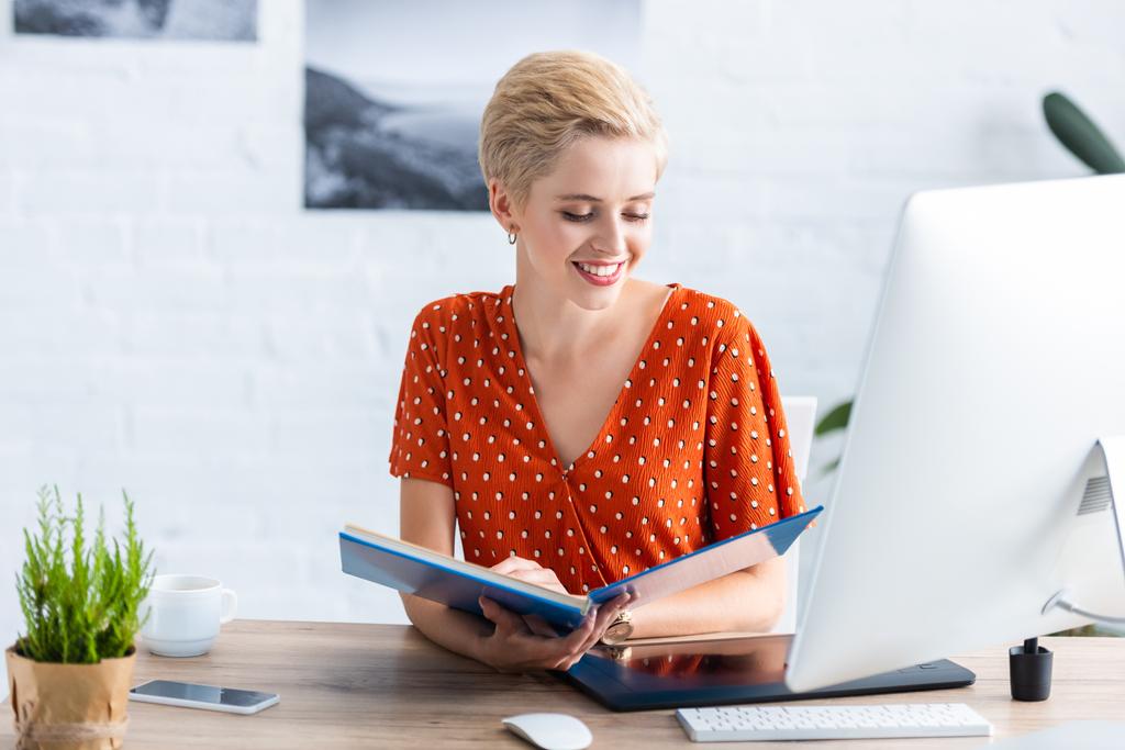 glimlachend vrouwelijke freelancer leesboek aan tafel met grafisch tablet en computer in kantoor aan huis  - Foto, afbeelding