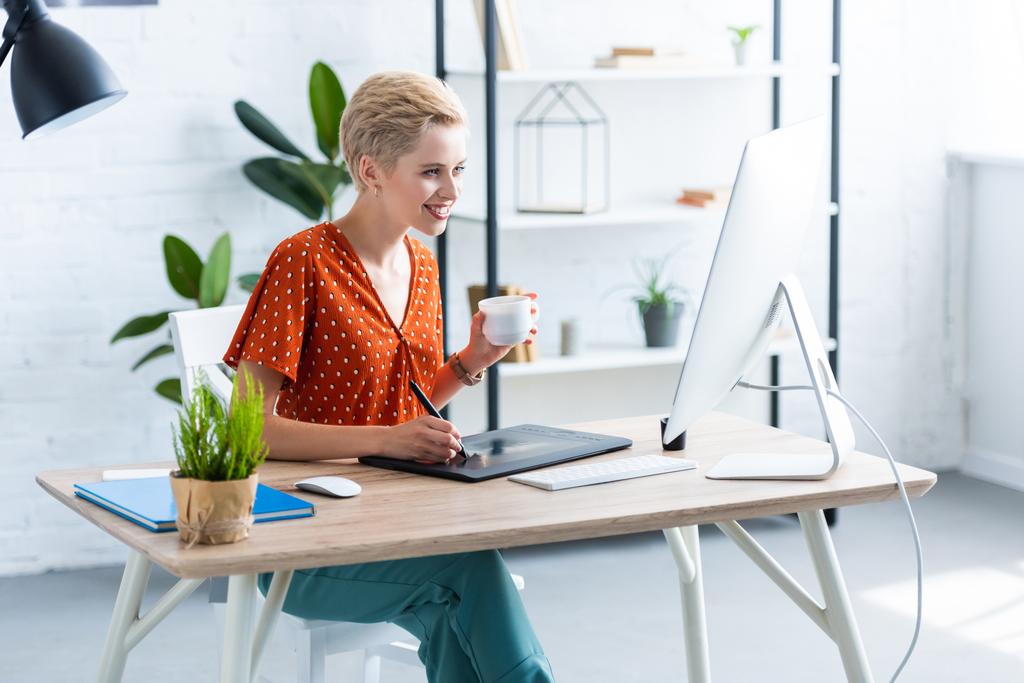 usmívající se žena na volné noze s šálek kávy na grafický tablet u stolu s počítačem  - Fotografie, Obrázek