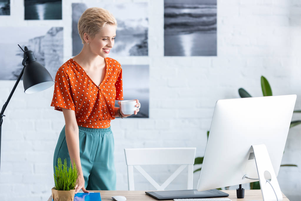 hymyilevä nainen freelancer tilalla kuppi kahvia lähellä pöytää graafinen tabletti ja tietokone kotitoimistossa
  - Valokuva, kuva