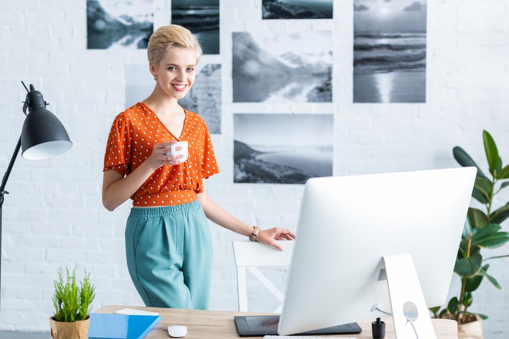 スタイリッシュな女性フリーランサー ホーム オフィスのグラフィック タブレットとコンピューターのテーブルの近くのコーヒーのカップを保持しています。  - 写真・画像