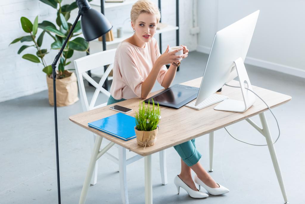 vista de ángulo alto del freelancer femenino sosteniendo taza de café en la mesa con tableta gráfica y computadora en la oficina en casa
  - Foto, Imagen