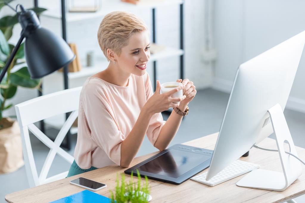 uśmiecha się kobieta freelancer picie kawy przy stole z tabletem graficznym i komputer w domu  - Zdjęcie, obraz