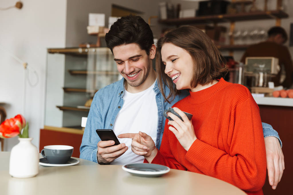 glückliches junges Paar sitzt am Cafétisch, trinkt Kaffee und schaut aufs Handy - Foto, Bild
