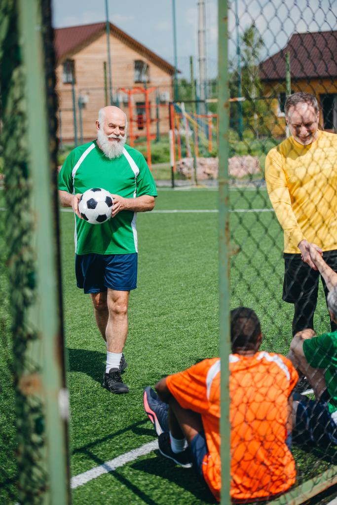 緑のフィールドで試合後に握手の異人種間の高齢者のサッカー選手 - 写真・画像