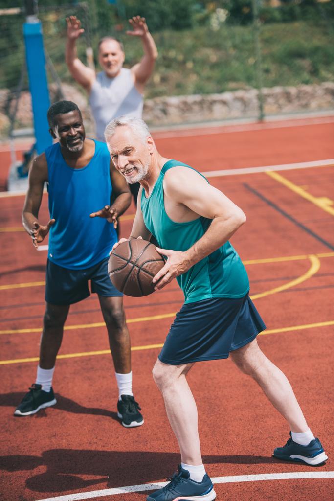 селективное внимание старика с баскетбольным мячом в руках и многорасовыми друзьями позади на детской площадке
 - Фото, изображение