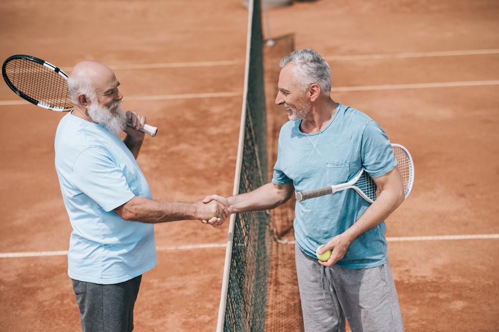 вид сбоку улыбающихся пожилых друзей с теннисными ракетками, пожимающими руку после игры на корте
 - Фото, изображение