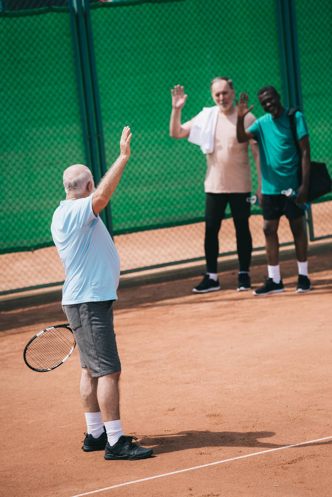 multiethnic old men greeting friend with tennis racquet on court - Photo, Image