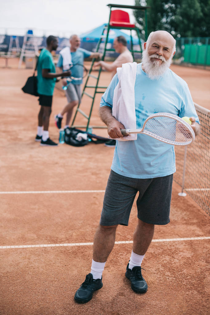 επιλεκτική εστίαση του ηλικιωμένος γενειοφόρος άνδρας με πετσέτα και το τένις ρακέτα βλέπουν φωτογραφική μηχανή, ενώ διαφυλετικός φίλοι έχοντας συνομιλία στο γήπεδο - Φωτογραφία, εικόνα