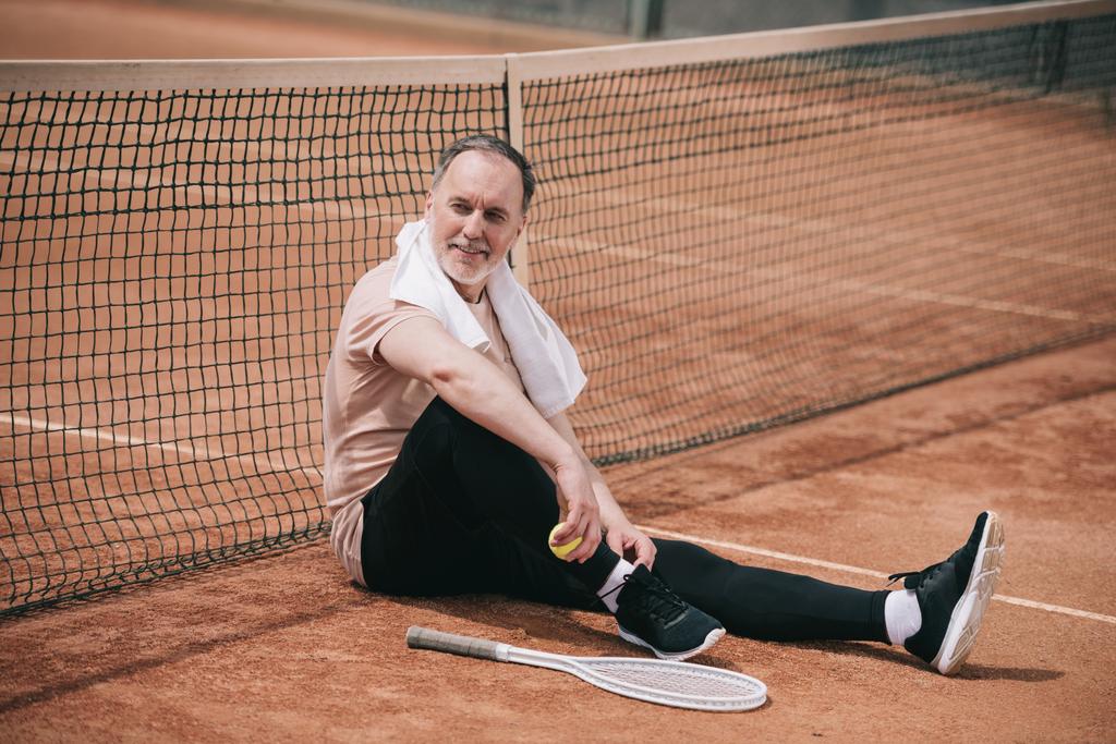 улыбающийся пожилой человек с полотенцем и теннисным оборудованием, отдыхающий возле сети на теннисном корте
 - Фото, изображение