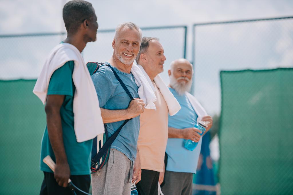 oldalnézetből a multikulturális idős férfiak, törölközőkkel és tenisz felszerelés a teniszpálya - Fotó, kép
