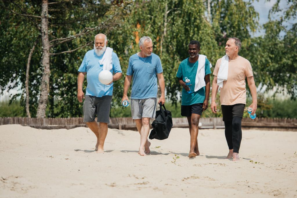 jugadores multiétnicos de voleibol de edad avanzada con botellas de agua deportiva caminando en la playa después del juego
 - Foto, imagen