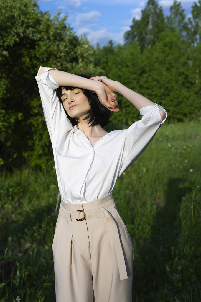Schöne stylische Mädchen tragen ein trendiges weißes Hemd, beige Hose und eine schwarze Perücke posieren entspannt im Freien im Sonnenlicht auf der grünen Wiese. Werbung und Modedesign. Kopierraum. - Foto, Bild