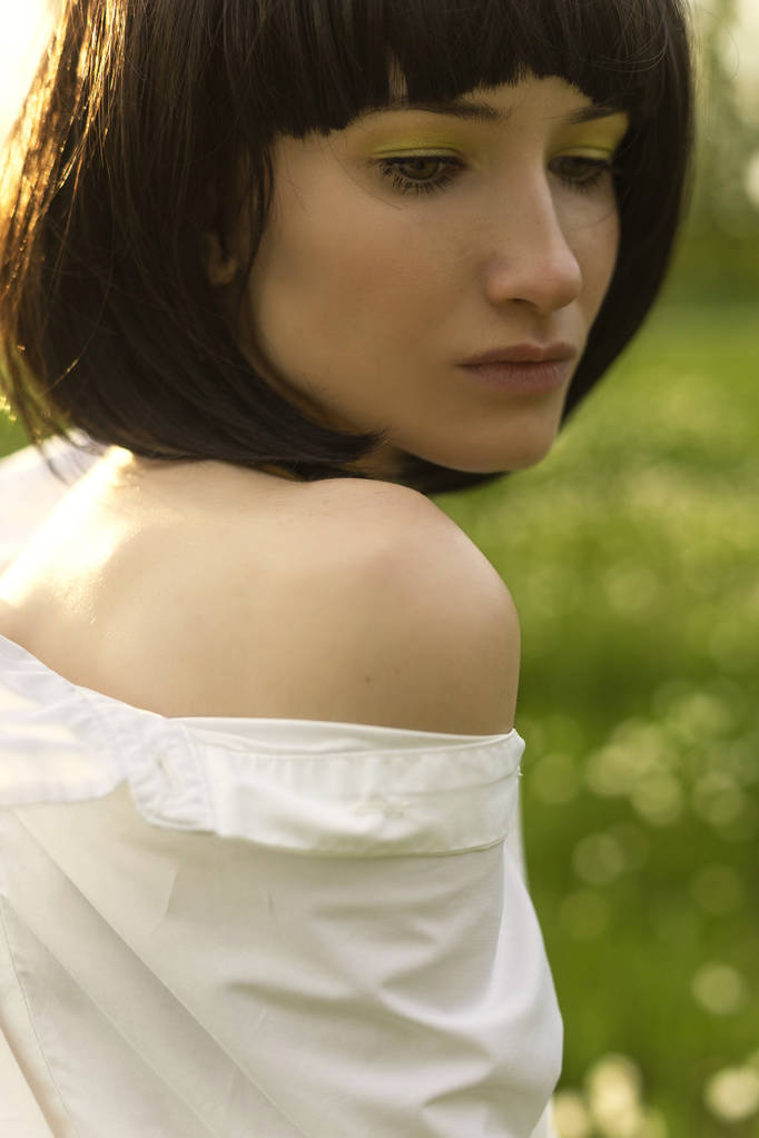 Στυλ art κοντά επάνω φωτογραφία ενός κοριτσιού με γυμνό ώμο φορώντας ένα μοντέρνο λευκό μπλουζάκι και μια μαύρη περούκα στο ηλιοβασίλεμα με φόντο πράσινο λιβάδι. Σχεδιασμός διαφήμισης και της μόδας. Χώρο αντίγραφο - Φωτογραφία, εικόνα