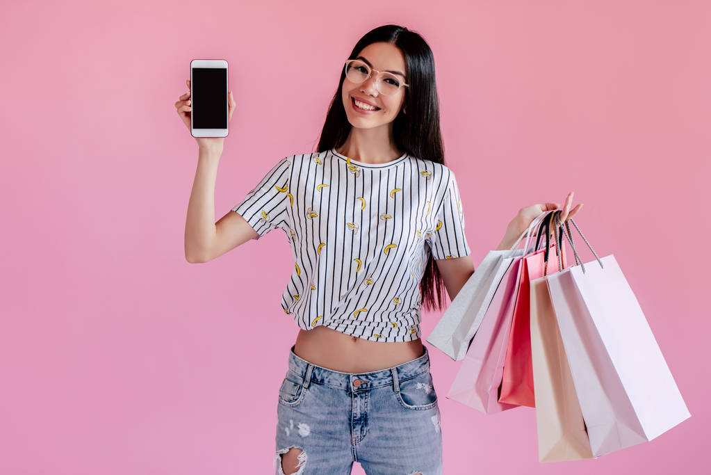 Adolescente asiática sobre fondo rosa. Elegante mujer joven con bolsas de compras y teléfono inteligente en las manos
 - Foto, Imagen